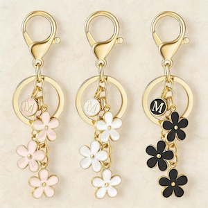 Könnte beinhalten: Drei goldfarbene Schlüsselanhänger mit Blumenanhängern. Jeder Schlüsselanhänger hat einen runden Verschluss, einen Ring und einen Anhänger mit dem Buchstaben "M". Die Blumenanhänger sind rosa, weiß und schwarz, mit goldfarbenen Kettengliedern.