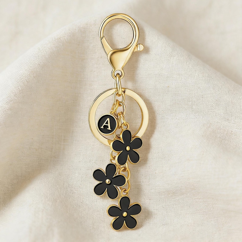 May include: Gold-tone keychain with a lobster clasp, a round charm with the letter "A", and three black flower charms. The keychain is hanging on a white fabric background.
