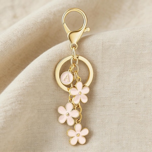 May include: Gold-colored keychain with a lobster clasp, a round ring, and a charm with the letter "A". Three pink flower charms dangle below. The keychain is a decorative accessory.