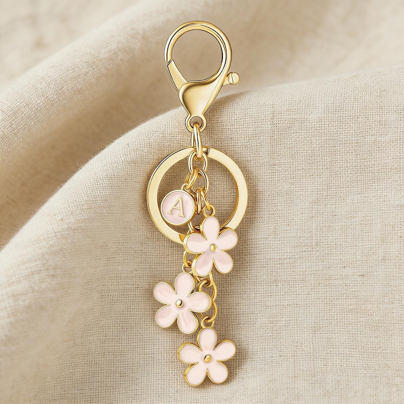 May include: Gold-colored keychain with a lobster clasp, a round ring, and a charm with the letter "A". Three pink flower charms dangle below. The keychain is a decorative accessory.