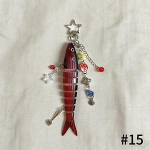 Puede incluir: Un llavero con forma de pez rojo y negro con un llavero plateado en forma de estrella. El llavero est&aacute; adornado con varios dijes, incluyendo estrellas, cuentas y un tenedor. El n&uacute;mero "#15" es visible.