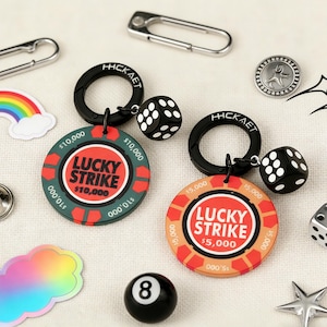 Può includere: Una collezione di accessori tra cui due portachiavi a dadi neri, due portachiavi a gettoni da poker con la scritta "LUCKY STRIKE", una palla 8 nera e vari ciondoli in metallo. I gettoni da poker sono rossi e verdi.