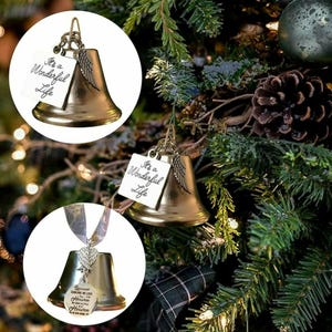 May include: Gold-colored bell Christmas ornaments with a wing charm and a white tag that reads "It's a Wonderful Life." The ornaments are hanging on a Christmas tree with other decorations.