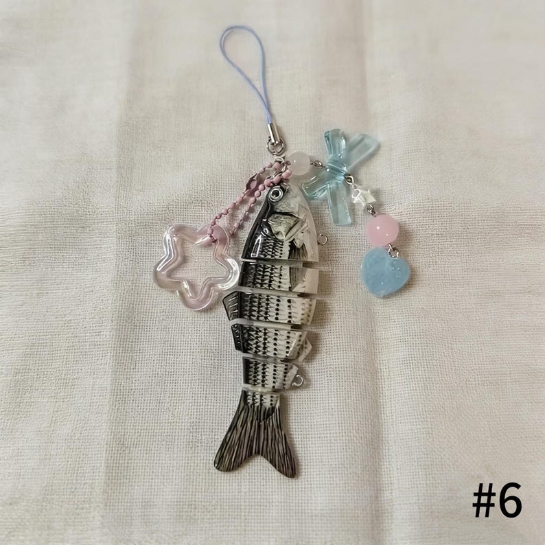 Puede incluir: Un dije con forma de pez con cuerpo transparente y detalles en blanco y negro. El dije est&aacute; sujeto a una cuerda azul claro y presenta una estrella rosa, un lazo azul y cuentas rosas y azules en forma de coraz&oacute;n. El n&uacute;mero 6 est&aacute; en la esquina inferior derecha.