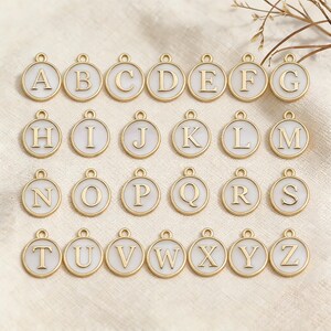 May include: Gold-colored alphabet charms, each with a white enamel background. The letters A through Z are displayed in a gold serif font. Each charm has a small loop at the top for attaching to a chain or bracelet. The charms are round.