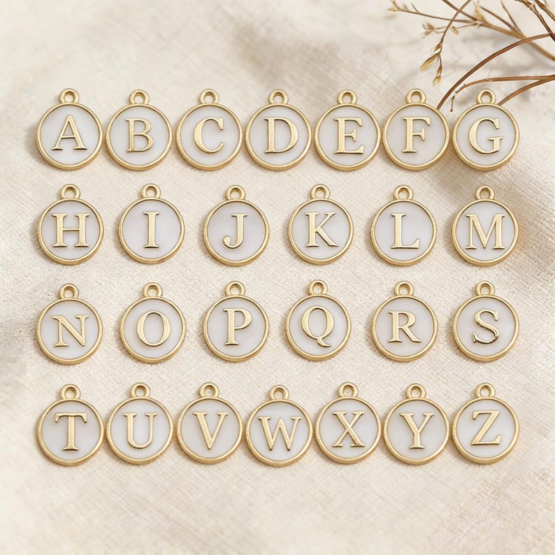 May include: Gold-colored alphabet charms, each with a white enamel background. The letters A through Z are displayed in a gold serif font. Each charm has a small loop at the top for attaching to a chain or bracelet. The charms are round.