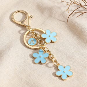 May include: Gold-tone keychain with a lobster clasp, a round charm with the letter "A", and three light blue flower charms. The flowers have a gold outline and a small gold dot in the center. The keychain is on a beige surface.