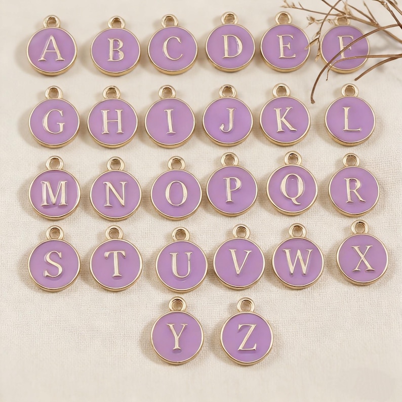 May include: A collection of 26 round charms, each featuring a different letter of the alphabet from A to Z. The charms have a light purple enamel background with a gold-colored border. Each letter is in a white, sans-serif font.