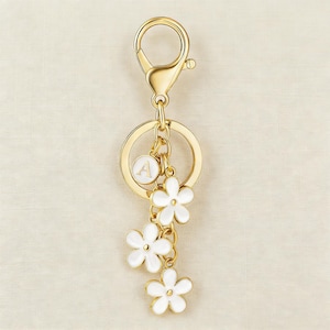 May include: Gold-tone keychain with a lobster clasp, a round charm with the letter "A", and three white flower charms. The keychain is a decorative accessory.