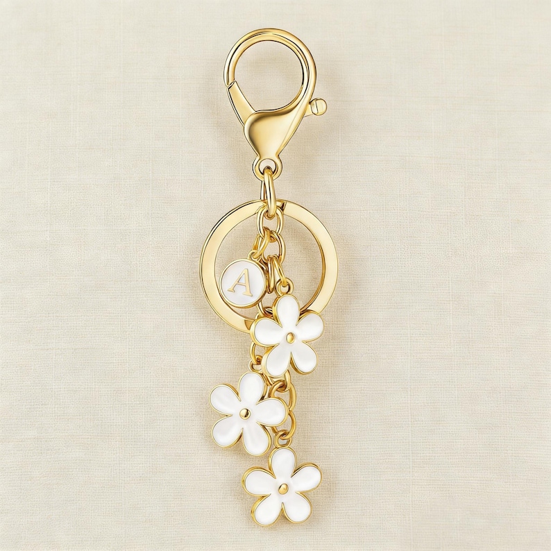 May include: Gold-tone keychain with a lobster clasp, a round charm with the letter "A", and three white flower charms. The keychain is a decorative accessory.