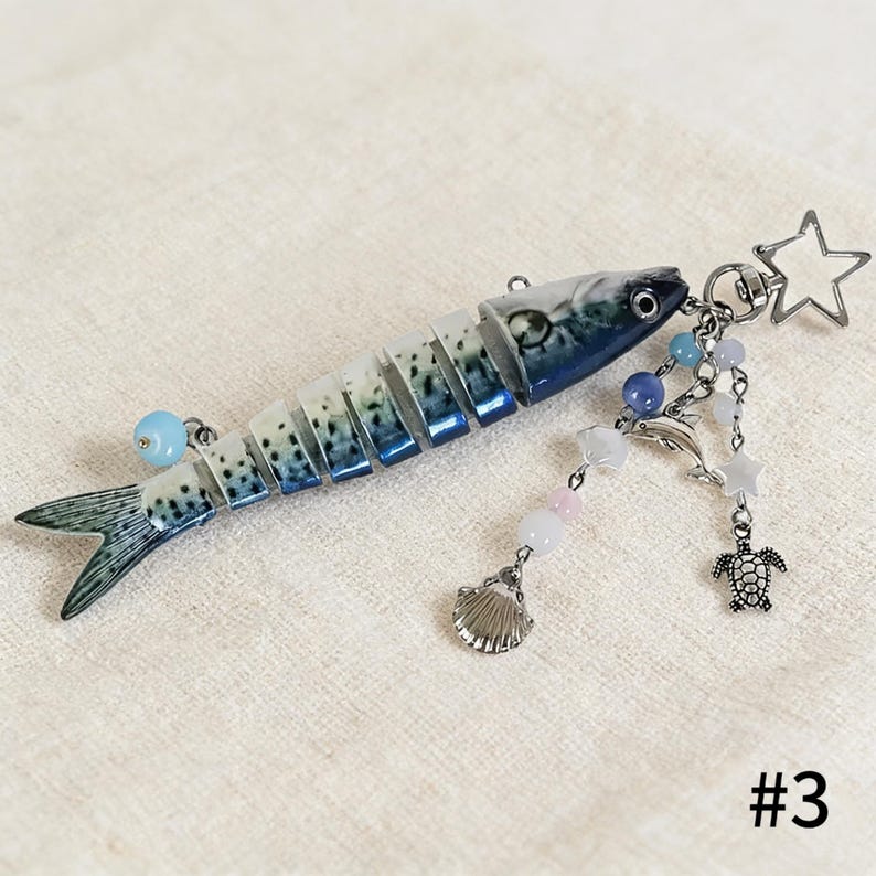 Puede incluir: Un llavero con forma de pez segmentado con detalles azules y blancos. El llavero tiene un dije de estrella, delf&iacute;n y tortuga plateados, junto con cuentas rosas y azules. El n&uacute;mero "#3" es visible en la esquina inferior derecha.