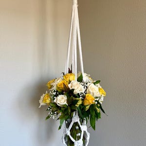 Può includere: Un portapiante in macramè bianco realizzato con corda annodata che sospende un vaso in vetro trasparente riempito con un bouquet di rose gialle e bianche, fogliame verde e piccoli fiori bianchi di gipsofila. Il supporto ha una lunga nappa.