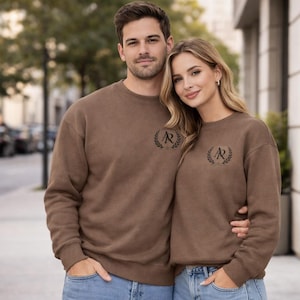 Puede incluir: Dos sudaderas marrones con la letra "R" dentro de un diseño de corona de laurel. Las sudaderas son de color marrón liso y parecen estar hechas de un material suave. Las sudaderas las lleva una pareja.