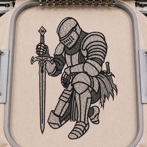 May include: An embroidery design on light beige fabric featuring a knight in full plate armor kneeling while holding a long sword upright. The knight is rendered in shades of gray and black thread with detailed outlines, secured within a gray embroidery hoop.
