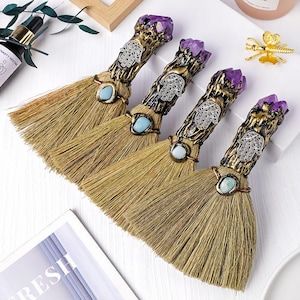 以下が含まれることがあります： 天然の毛を使った4つの装飾用ほうき。各ほうきは、紫色のクリスタル、ターコイズ色の石、銀色のハムサハンドデザインで飾られたブロンズ色のハンドルが特徴です。ほうきは白い表面に配置されています。