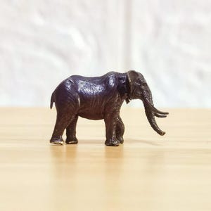 May include: A small, dark brown elephant figurine with a textured surface. The elephant has a long trunk, large ears, and visible tusks. The figurine is standing on a light-colored wooden surface.
