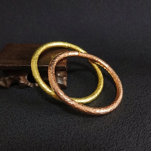 May include: Two hammered metal bangles. One is gold-coloured, the other is copper-coloured. The bangles are circular and have a textured surface. They are displayed on a dark surface.