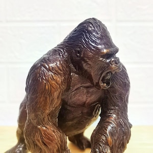Può includere: Una statuetta di gorilla marrone scuro con una finitura in pelliccia testurizzata e una muscolatura dettagliata. Il gorilla è raffigurato in una posizione inclinata in avanti con la bocca aperta, che mostra i denti, e la testa girata.