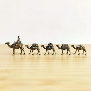 Könnte beinhalten: Eine Reihe von fünf kleinen, bronzefarbenen Kamel-Figuren. Das erste Kamel hat einen Reiter, die anderen tragen Packungen. Die Figuren stehen auf einer hellen Holzoberfläche, mit einem schlichten weißen Hintergrund.
