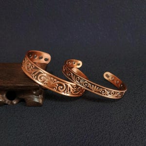 Könnte beinhalten: Zwei kupferfarbene Armreifen mit aufwendigen floralen und scrollartigen Designs. Die offenen Armreifen werden auf einer dunklen Oberfläche präsentiert, die ihre detaillierten Muster und warmen metallischen Töne hervorhebt. Vintage-Ästhetik.