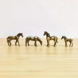 May include: Four small, bronze-coloured horse figurines are arranged on a light-coloured wooden surface. The horses are in various poses, including standing and grazing. The smallest horse is a foal.