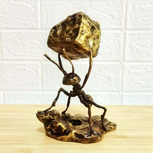 May include: A bronze-toned metal sculpture of an ant standing on a textured base, holding a large, pitted, rock-like object above its head with its front legs. The figure features a dark, metallic finish with highlights on the raised surfaces.