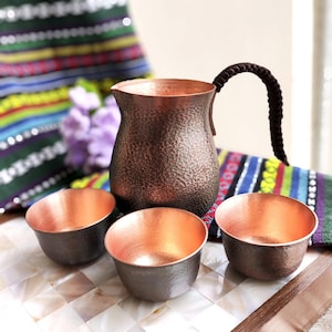 May include: A hammered copper pitcher with a dark brown braided handle and three matching hammered copper cups. The pitcher has a wide base and a narrow neck, while the cups feature a smooth, polished interior and a textured exterior finish.