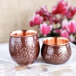 May include: Two copper-coloured, round bowls with embossed floral designs. The bowls have a hammered texture and a shiny, polished interior. The bowls are on a white, patterned surface, with pink flowers in the background.