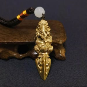 May include: A gold-toned metal pendant featuring a detailed Ganesha figure seated on a lotus flower, positioned above a pointed vajra-style base. The pendant is attached to a dark cord with a single white bead and decorative yellow and red accents.