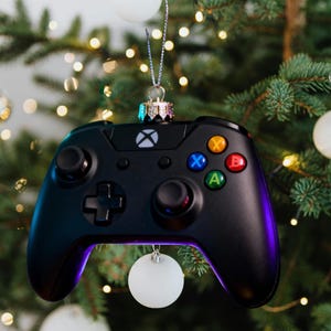May include: Black Xbox controller Christmas tree ornament. The controller has coloured buttons and a silver hanging loop. The tree is decorated with white and gold baubles.