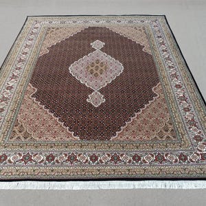 May include: A rectangular rug with an intricate pattern. The central design is a diamond shape in shades of brown, beige, and pink. The border features a repeating floral pattern with a black edge and white fringe.