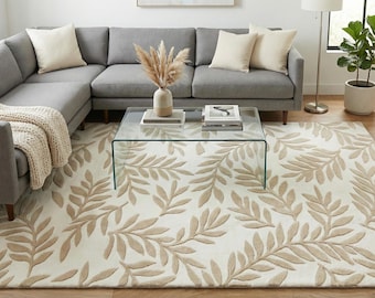 May include: A large, rectangular area rug with a cream-colored background and a repeating pattern of beige-colored leaves. The rug is placed in a living room setting, under a glass coffee table and a gray sectional sofa.