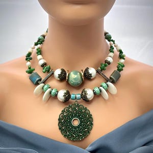 Puede incluir: Collar de dos hilos con un gran colgante verde adornado. El collar incorpora cuentas blancas, verdes y azules, con una mezcla de formas y texturas. El collar se exhibe en un maniquí. El colgante tiene un agujero central.