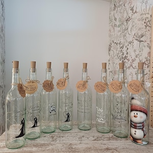 Könnte beinhalten: Eine Sammlung klarer Glasflaschen mit Korken, jede mit Lichterketten und dekorativen Anhängern versehen. Einige Flaschen weisen aufgedruckte Designs und Texte auf, darunter eine Schneemann-Illustration. Die Flaschen sind auf einer Holzoberfläche angeordnet.