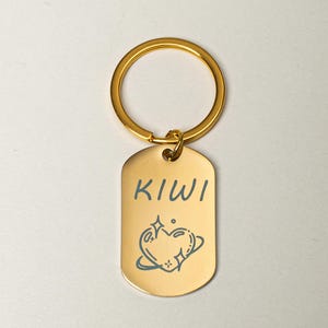 Puede incluir: Un llavero de metal dorado con una etiqueta rectangular. La etiqueta presenta la palabra "KIWI" en escritura azul sobre un diseño de corazón y planeta azul. El llavero tiene un anillo dorado para sujetar las llaves.