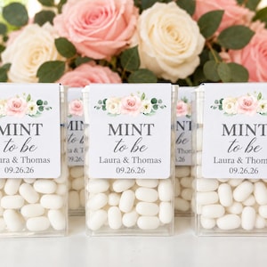 May include: Clear rectangular plastic containers filled with white mints, featuring a label with a floral illustration, the text MINT to be, the names Laura & Thomas, and the date 09.26.26, set against a background of pink and white roses.