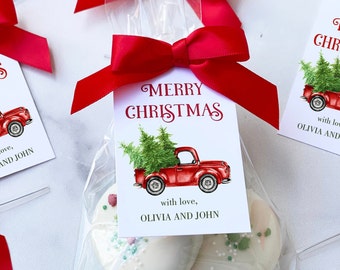 May include: Clear cellophane bag tied with a red ribbon, containing two white-frosted cookies. A tag with "MERRY CHRISTMAS" and a red truck carrying Christmas trees is attached. The tag also includes the text "with love, OLIVIA AND JOHN."