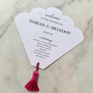 May include: A white wedding program with a scalloped edge, featuring the names Darian & Brandon and the date 09.27.25. The program lists the ceremony events and has a bright pink tassel attached.