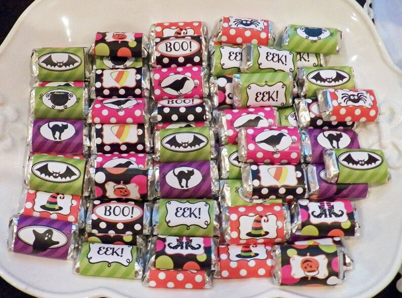 May include: A collection of Halloween-themed candy bars with colorful wrappers. The wrappers feature various designs, including bats, spiders, and the words "BOO!" and "EEK!" The candies are arranged on a white plate, creating a festive display.