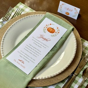 K&ouml;nnte beinhalten: Herbstliche Tischdekoration mit wei&szlig;em Teller, gr&uuml;ner Serviette und Tischkarte. Die Tischkarte und das Men&uuml; zeigen eine K&uuml;rbisillustration und das Wort "Grateful". Das Set beinhaltet einen Jutenteller, Besteck und eine gr&uuml;n-wei&szlig; karierte Tischdecke.