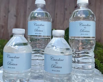 May include: Four clear plastic water bottles with white caps. Each bottle has a light blue label with the names "Caroline & Brian" and the date "09.02.2026" printed in a script font. The bottles are filled with water.