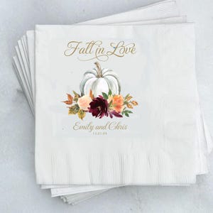 May include: White paper napkins with a printed design. The design features a white pumpkin, floral arrangement, and the text "Fall in Love," along with names and a date. The napkins are stacked, showing a textured surface.