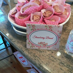 May include: A platter of strawberry bread cupcakes in pink and white paper liners. The cupcakes are arranged in a circle on a white platter with a pink and white floral design. A sign in the foreground reads "Strawberry Bread".