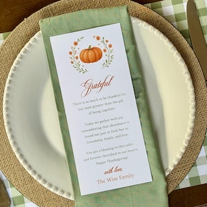K&ouml;nnte beinhalten: Ein Thanksgiving-Tischarrangement mit einem wei&szlig;en Teller, einer gr&uuml;nen Serviette und einer Karte mit dem Wort "Grateful". Die Karte zeigt einen Aquarellk&uuml;rbis und ein Blumendesign. Der Text dr&uuml;ckt Dankbarkeit und Thanksgiving-W&uuml;nsche aus.