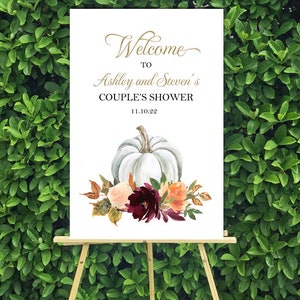 May include: A white sign on a wooden easel with the text "Welcome to Ashley and Steven's COUPLE'S SHOWER 11.10.22". The sign features a watercolor illustration of a white pumpkin and floral arrangement with orange, burgundy, and peach flowers.