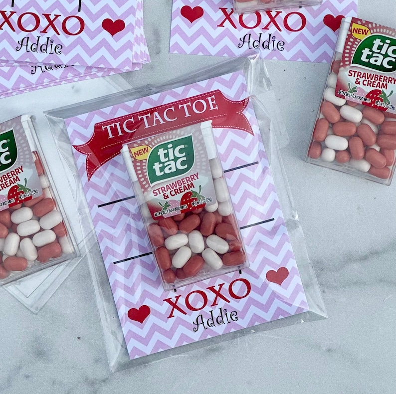 May include: A clear plastic bag with a pink and white chevron pattern card inside. The card has the words 'Tic Tac Toe' in red with a red ribbon. The card also has the words 'XOXO Addie' in purple with a pink heart. The bag contains two small boxes of Tic Tacs, one with the words 'New Tic Tac Strawberry & Cream' and the other with the words 'Strawberry & Cream'. The Tic Tacs are in a clear plastic container with a white lid.