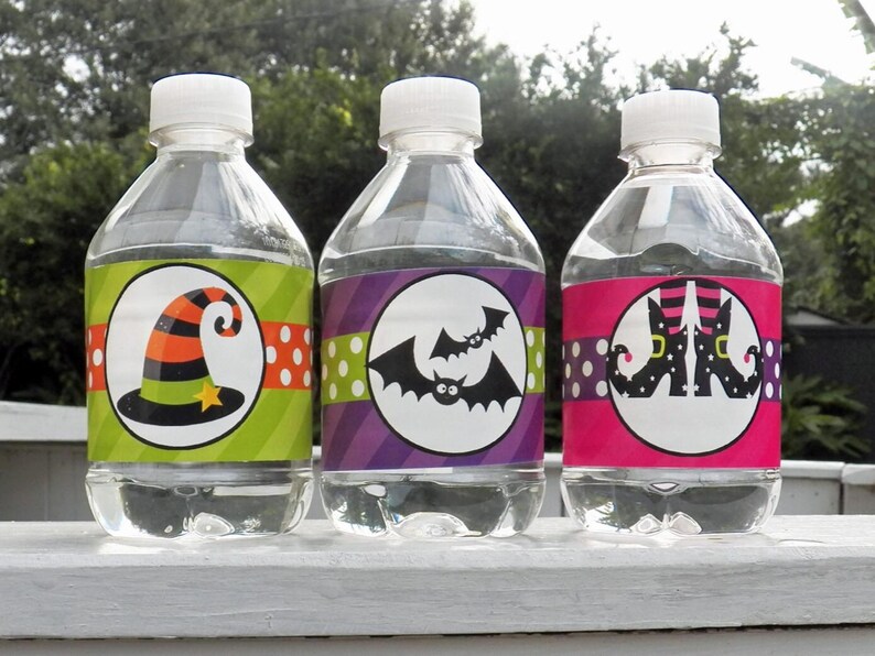 May include: Three clear water bottles with white caps, each featuring a Halloween-themed label. The labels display a witch's hat, bats, and witch's legs, with polka dot and striped backgrounds. The bottles are displayed outdoors.