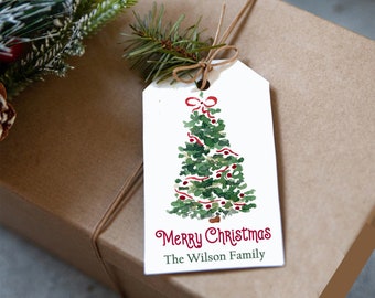 May include: A gift tag with a watercolor Christmas tree design, red ribbon, and the words "Merry Christmas" and "The Wilson Family" in red. The tag is tied to a wrapped brown gift box with twine and a sprig of evergreen.