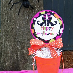 May include: A Halloween-themed display featuring a bright orange bucket with a polka-dot bow, filled with shredded paper. A sign with witch boots and the words "Happy Halloween" is displayed. A large black spider is attached to a tree.