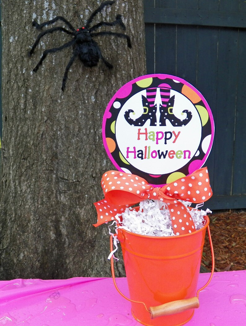 May include: A Halloween-themed display featuring a bright orange bucket with a polka-dot bow, filled with shredded paper. A sign with witch boots and the words "Happy Halloween" is displayed. A large black spider is attached to a tree.